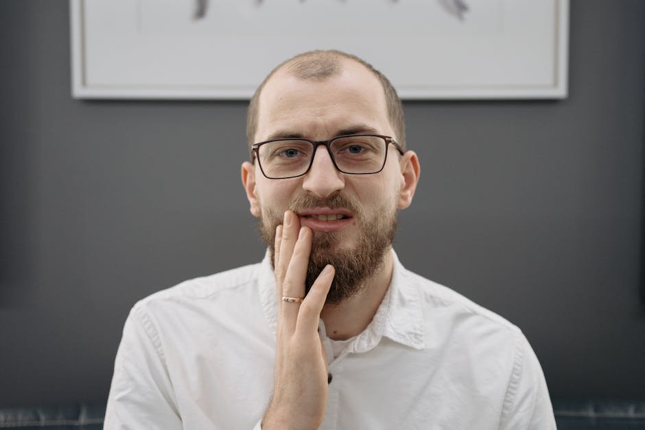 A man with eyeglasses and beard shows discomfort from a toothache indoors.