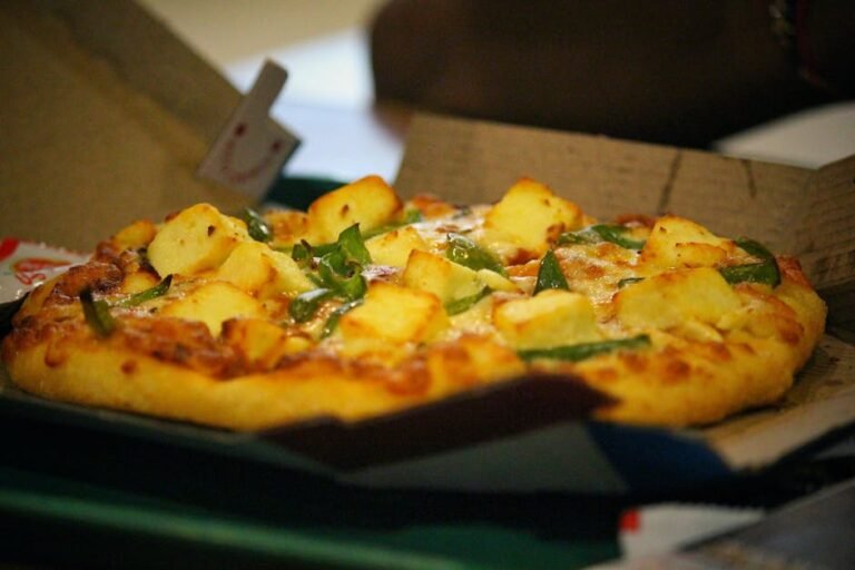 A delicious cheesy pizza topped with paneer and peppers, served in a cardboard box. Mouth-watering and ready to enjoy.